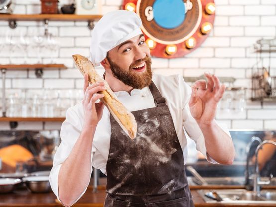 Panadero guapo feliz con barra de pan y mostrando signo de ok en la cocina